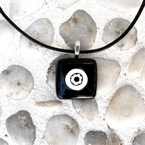 May include: A square black pendant with a white floral design, suspended from a black cord. The pendant has a silver-coloured bail. The background is composed of white and grey stones.