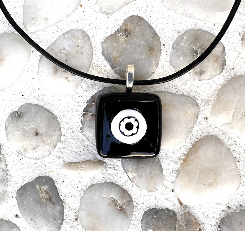 May include: A square black pendant with a white floral design, suspended from a black cord. The pendant has a silver-coloured bail. The background is composed of white and grey stones.