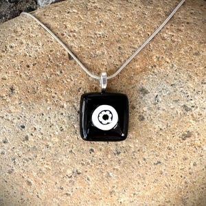May include: A square black pendant necklace with a white floral design. The pendant is attached to a silver chain. The necklace is displayed on a textured, light brown surface.