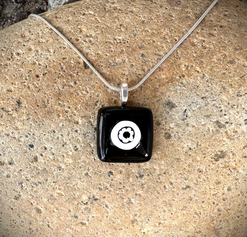 May include: A square black pendant necklace with a white floral design. The pendant is attached to a silver chain. The necklace is displayed on a textured, light brown surface.
