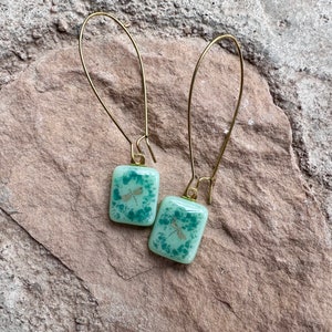 May include: A pair of gold-colored drop earrings. Each earring features a square, light green glass pendant with a dragonfly design. The earrings are set against a textured, light brown stone surface.