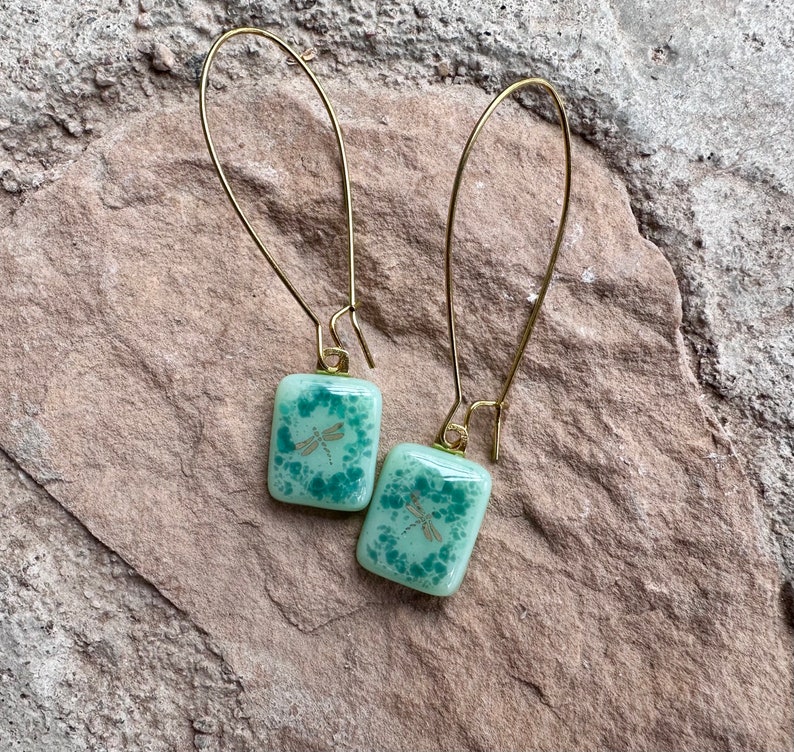 May include: A pair of gold-colored drop earrings. Each earring features a square, light green glass pendant with a dragonfly design. The earrings are set against a textured, light brown stone surface.