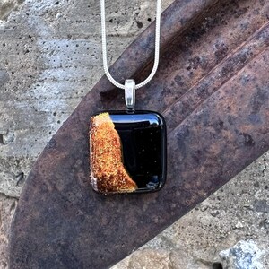 Puede incluir: Un collar con colgante cuadrado y cadena plateada. El colgante es negro con una secci&oacute;n texturizada de color marr&oacute;n dorado en el lado izquierdo. El colgante est&aacute; sujeto a una fianza plateada.