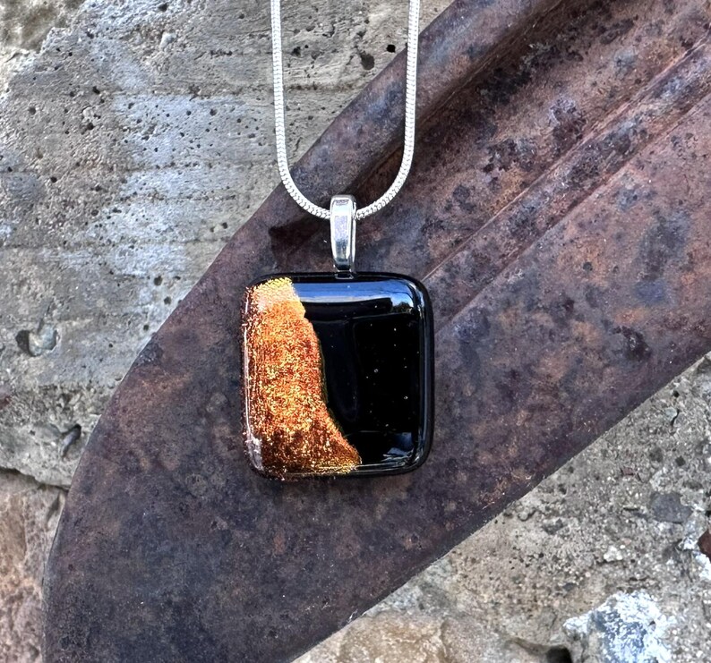 Puede incluir: Un collar con colgante cuadrado y cadena plateada. El colgante es negro con una secci&oacute;n texturizada de color marr&oacute;n dorado en el lado izquierdo. El colgante est&aacute; sujeto a una fianza plateada.