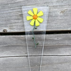 May include: A clear glass flower stake featuring a yellow daisy with an orange center. The flower has a green stem with small leaves. The stake is a clear, elongated shape, perfect for garden decor.