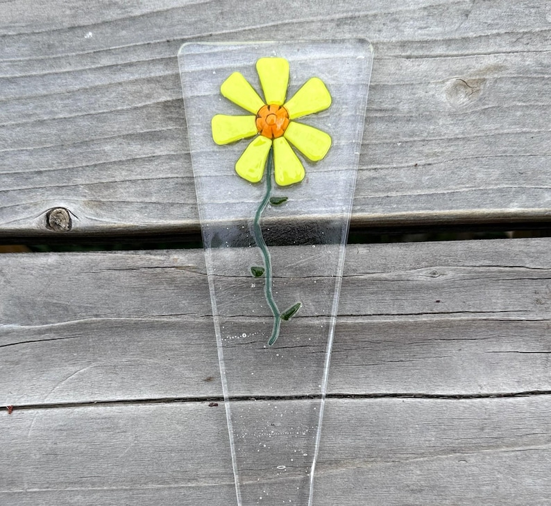 May include: A clear glass flower stake featuring a yellow daisy with an orange center. The flower has a green stem with small leaves. The stake is a clear, elongated shape, perfect for garden decor.