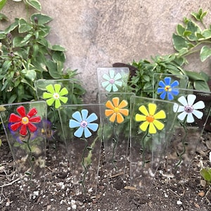 May include: A collection of colorful glass flower garden stakes. Each stake features a different colored flower, including red, blue, yellow, orange, green, and white. The stakes are displayed in a garden setting, with soil and greenery in the background.
