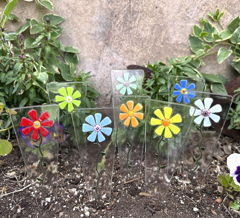 May include: A collection of colorful glass flower garden stakes. Each stake features a different colored flower, including red, blue, yellow, orange, green, and white. The stakes are displayed in a garden setting, with soil and greenery in the background.