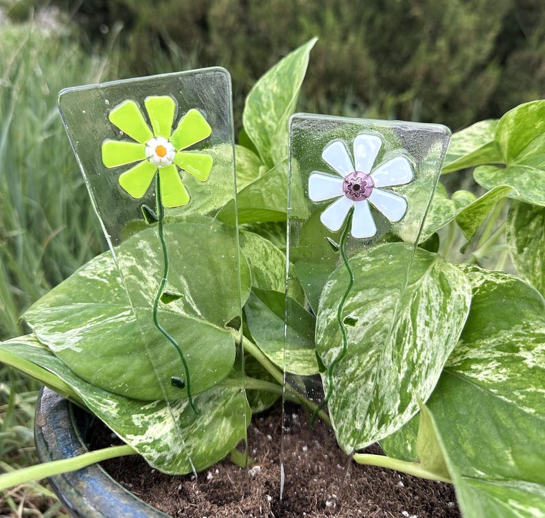 May include: Two glass flower plant stakes. One stake features a light green flower with a white and orange center. The other stake has a white flower with a purple center. Both have green stems and are set in a potted plant.