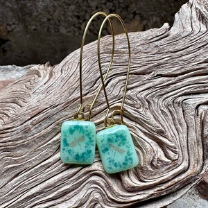 May include: A pair of handmade earrings featuring square, light green glass pendants with a dragonfly design. The pendants are suspended from gold-colored, long wire hooks. The earrings are displayed on a piece of weathered wood.