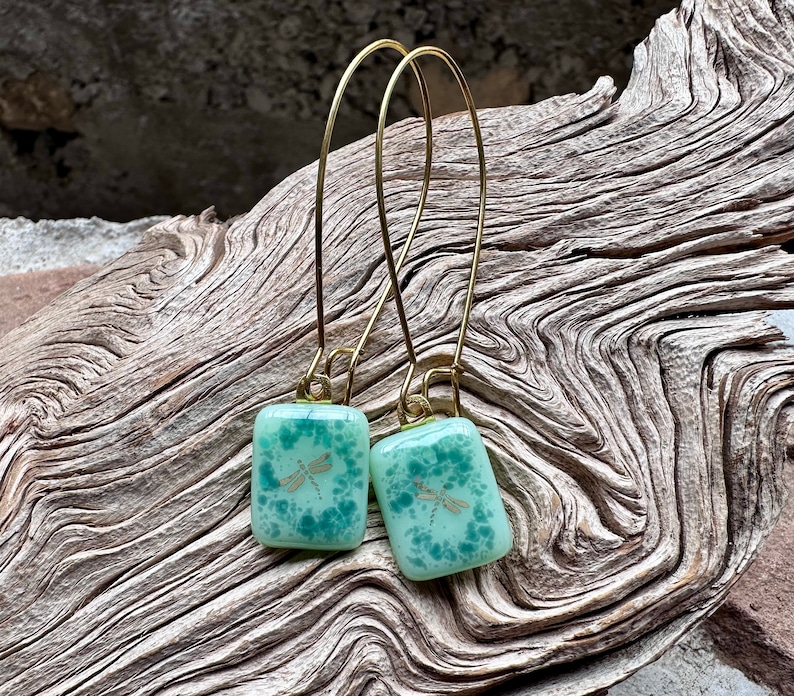 May include: A pair of handmade earrings featuring square, light green glass pendants with a dragonfly design. The pendants are suspended from gold-colored, long wire hooks. The earrings are displayed on a piece of weathered wood.