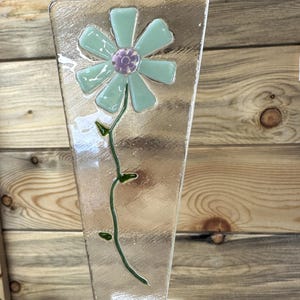 May include: A clear glass decorative piece with a flower design. The flower has light blue petals and a purple center. A green stem with leaves extends down the glass. The glass has a textured surface.