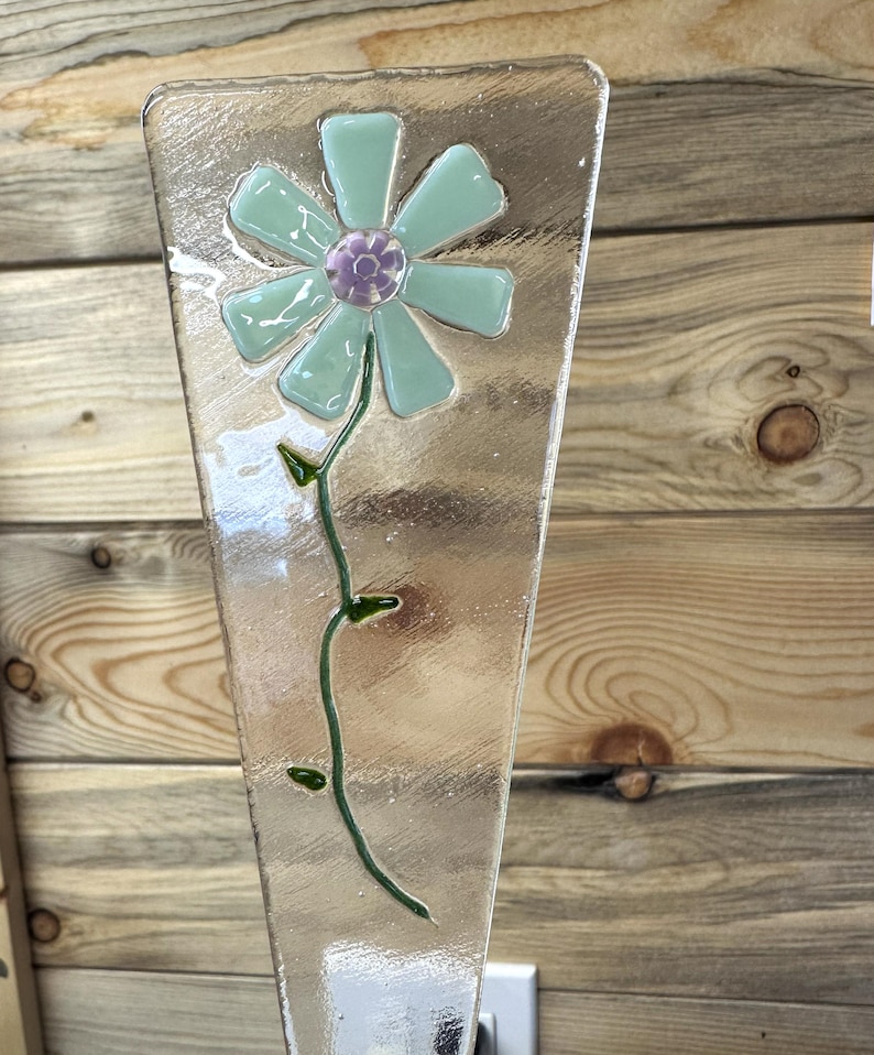 May include: A clear glass decorative piece with a flower design. The flower has light blue petals and a purple center. A green stem with leaves extends down the glass. The glass has a textured surface.