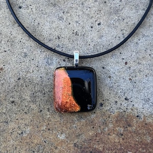 Puede incluir: Un collar con colgante cuadrado con fondo negro y una secci&oacute;n de vidrio fundido naranja y dorado. El colgante est&aacute; sujeto a un cord&oacute;n negro con una fianza de color plateado.