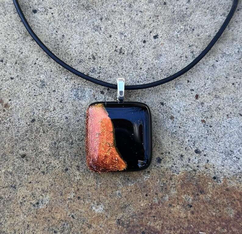Puede incluir: Un collar con colgante cuadrado con fondo negro y una secci&oacute;n de vidrio fundido naranja y dorado. El colgante est&aacute; sujeto a un cord&oacute;n negro con una fianza de color plateado.
