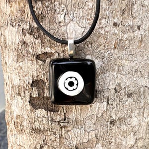 May include: A black square pendant necklace featuring a white floral design. The pendant is suspended from a black cord with a silver-coloured bail. The necklace is set against a textured, brown tree trunk.