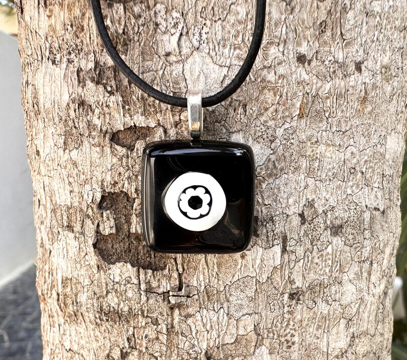 May include: A black square pendant necklace featuring a white floral design. The pendant is suspended from a black cord with a silver-coloured bail. The necklace is set against a textured, brown tree trunk.