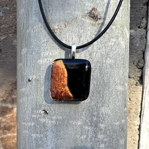 Puede incluir: Un collar con colgante cuadrado con un dise&ntilde;o de vidrio negro y color cobre. El colgante est&aacute; suspendido de un cord&oacute;n negro con un cierre plateado. El vidrio color cobre tiene una apariencia texturizada y brillante.
