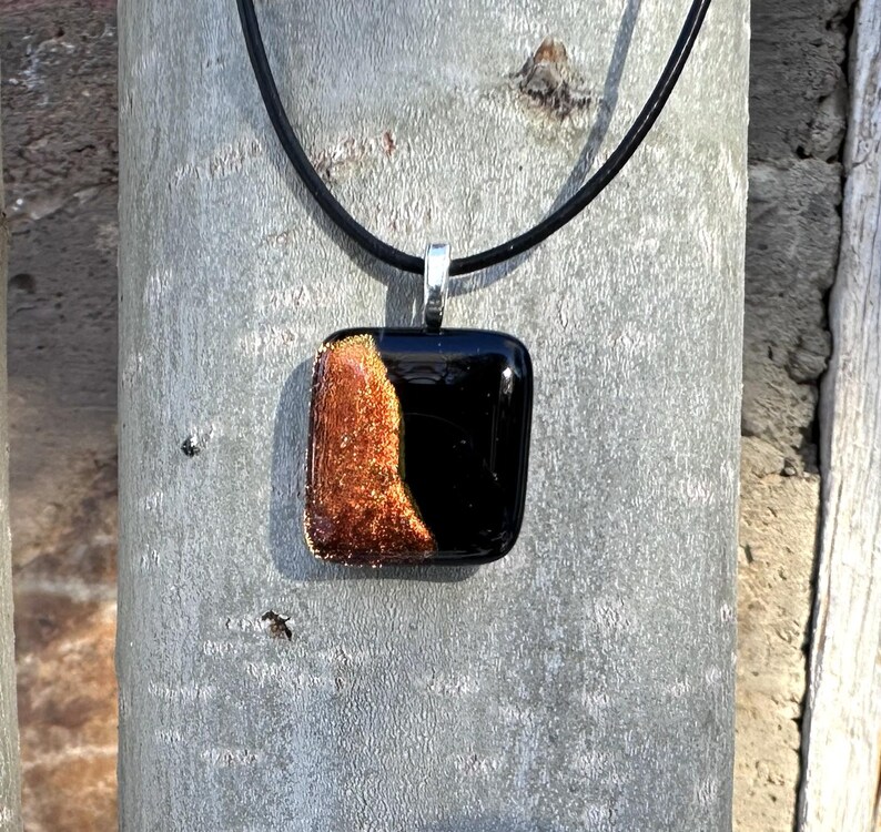 Puede incluir: Un collar con colgante cuadrado con un dise&ntilde;o de vidrio negro y color cobre. El colgante est&aacute; suspendido de un cord&oacute;n negro con un cierre plateado. El vidrio color cobre tiene una apariencia texturizada y brillante.
