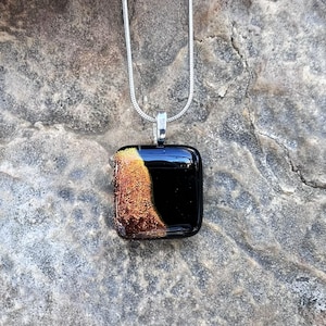 Puede incluir: Un collar con colgante cuadrado con un dise&ntilde;o negro y dorado. El colgante es negro brillante con una secci&oacute;n dorada texturizada en el lado izquierdo. La cadena del collar es plateada.