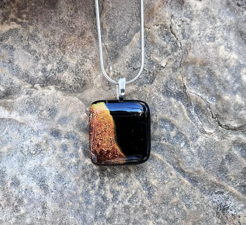 Puede incluir: Un collar con colgante cuadrado con un dise&ntilde;o negro y dorado. El colgante es negro brillante con una secci&oacute;n dorada texturizada en el lado izquierdo. La cadena del collar es plateada.