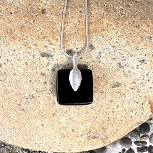 May include: A silver necklace featuring a square, black pendant. A silver leaf-shaped charm is attached to the pendant. The necklace chain is a thin, silver snake chain. The jewellery is displayed on a textured, tan stone surface.