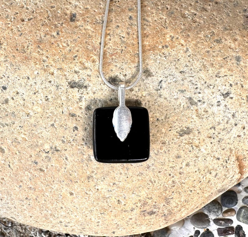 May include: A silver necklace featuring a square, black pendant. A silver leaf-shaped charm is attached to the pendant. The necklace chain is a thin, silver snake chain. The jewellery is displayed on a textured, tan stone surface.