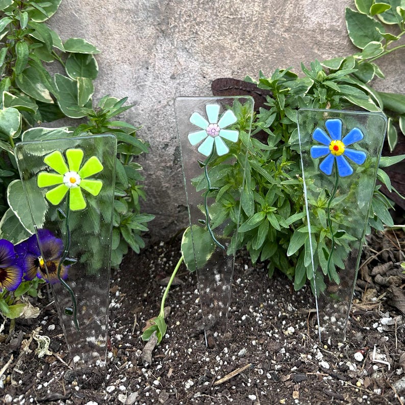 May include: Three clear glass garden stakes with colorful flower designs. The flowers are yellow, light blue, and blue with yellow centers. The stakes are placed in soil among green plants, creating a decorative garden accent.