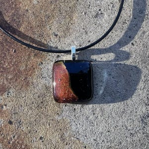 Puede incluir: Un collar con colgante cuadrado con un cord&oacute;n de cuero negro. El colgante tiene forma rectangular con una fianza plateada. El colgante tiene una combinaci&oacute;n de colores negro y marr&oacute;n con motas doradas.