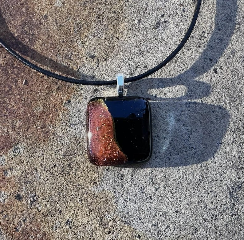 Puede incluir: Un collar con colgante cuadrado con un cord&oacute;n de cuero negro. El colgante tiene forma rectangular con una fianza plateada. El colgante tiene una combinaci&oacute;n de colores negro y marr&oacute;n con motas doradas.