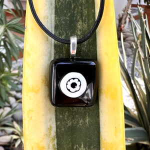 May include: A black square pendant with a white floral design, suspended from a black cord. The pendant has a silver-coloured bail. The background features a yellow and green plant.