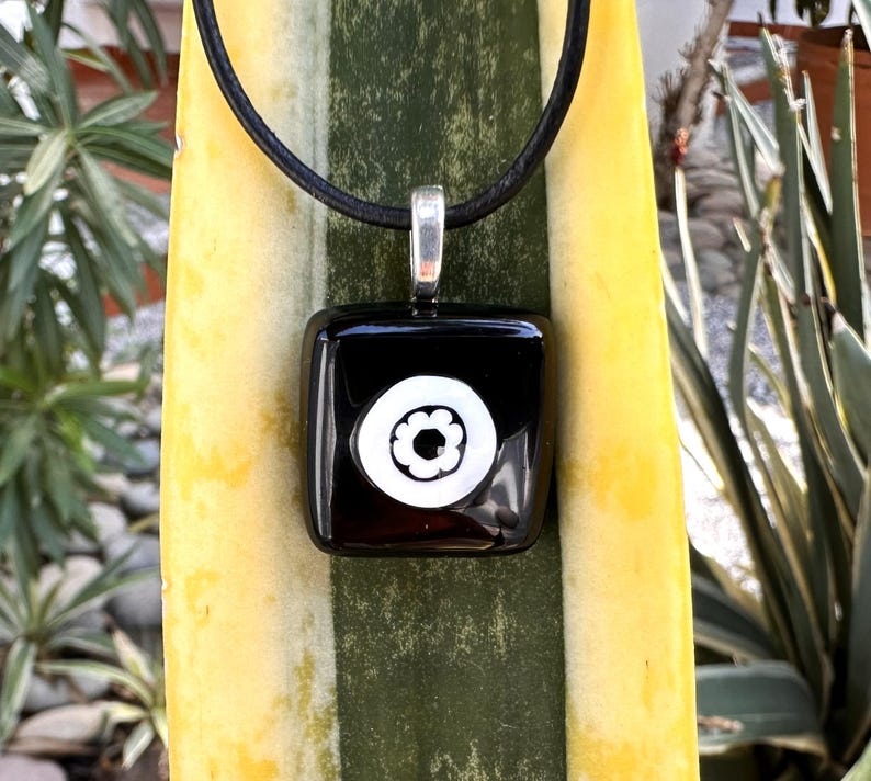May include: A black square pendant with a white floral design, suspended from a black cord. The pendant has a silver-coloured bail. The background features a yellow and green plant.