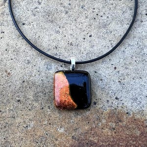 Puede incluir: Un collar con colgante cuadrado de vidrio negro y cobrizo. El colgante est&aacute; sujeto a un collar de cord&oacute;n negro con un cierre plateado. El colgante tiene una superficie brillante y reflectante.