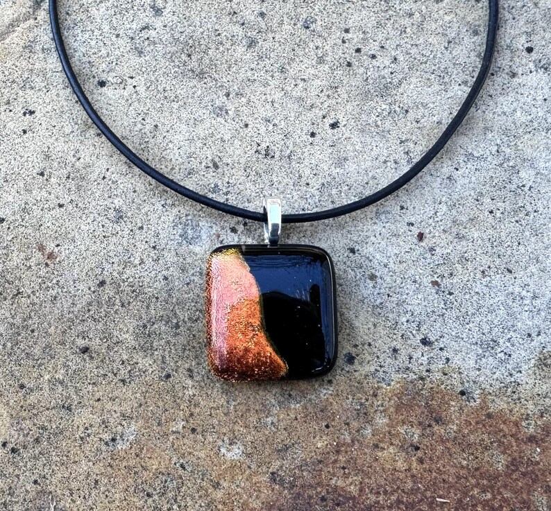 Puede incluir: Un collar con colgante cuadrado de vidrio negro y cobrizo. El colgante est&aacute; sujeto a un collar de cord&oacute;n negro con un cierre plateado. El colgante tiene una superficie brillante y reflectante.