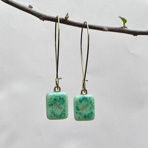 May include: A pair of dangle earrings featuring square, light green glass pendants with a dragonfly design. The earrings have gold-colored, long, thin hooks. The glass has a speckled pattern, and the dragonfly is a light gold color.