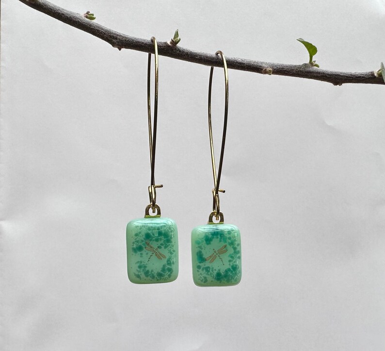 May include: A pair of dangle earrings featuring square, light green glass pendants with a dragonfly design. The earrings have gold-colored, long, thin hooks. The glass has a speckled pattern, and the dragonfly is a light gold color.