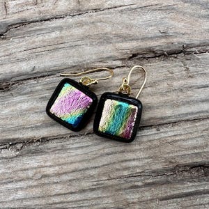 May include: A pair of square earrings with iridescent glass and gold accents. The earrings are on a wooden surface.