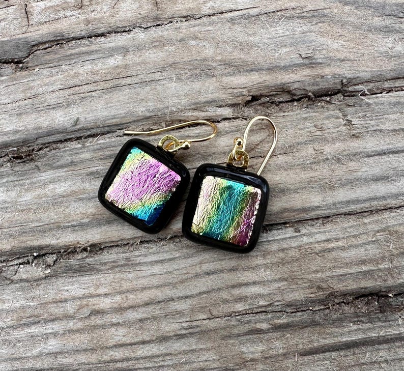 May include: A pair of square earrings with iridescent glass and gold accents. The earrings are on a wooden surface.