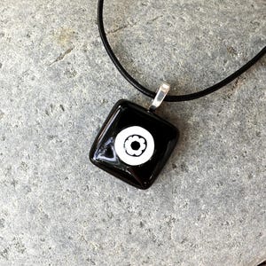 May include: Black square pendant necklace featuring a white floral design. The pendant hangs from a black cord with a silver-coloured bail. The necklace is shown on a grey stone surface.