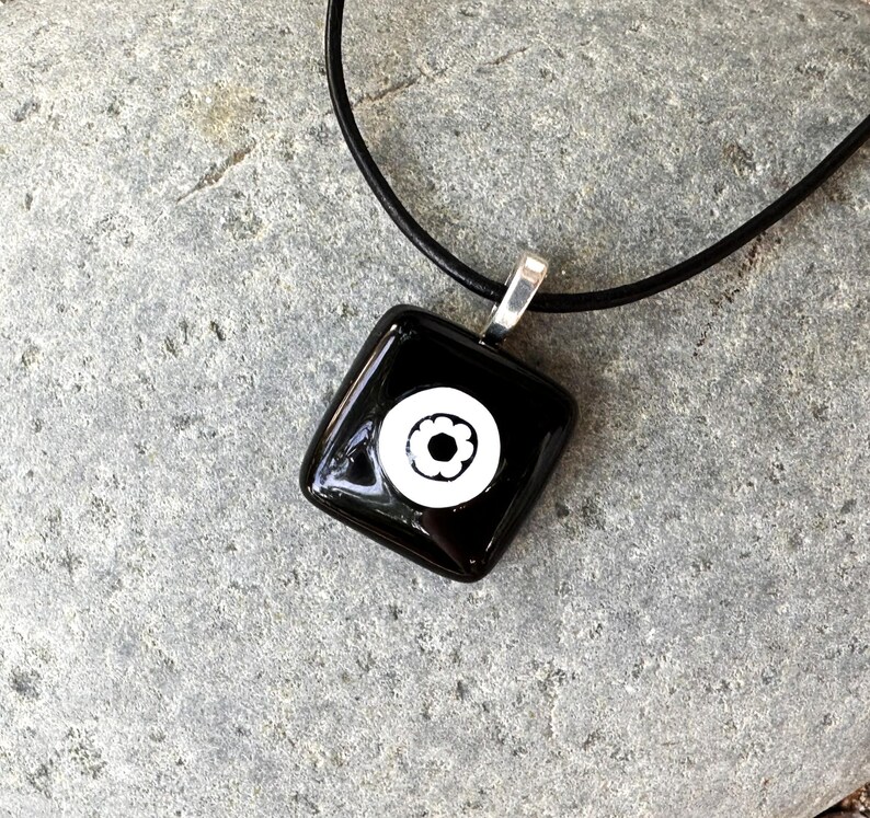 May include: Black square pendant necklace featuring a white floral design. The pendant hangs from a black cord with a silver-coloured bail. The necklace is shown on a grey stone surface.