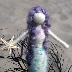 May include: A handmade felt mermaid doll with long purple hair, a white body, and a blue and white patterned tail. The doll is holding a white starfish.