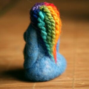 May include: A blue felt figurine with a rainbow yarn hair. The figurine is sitting on a wooden surface.