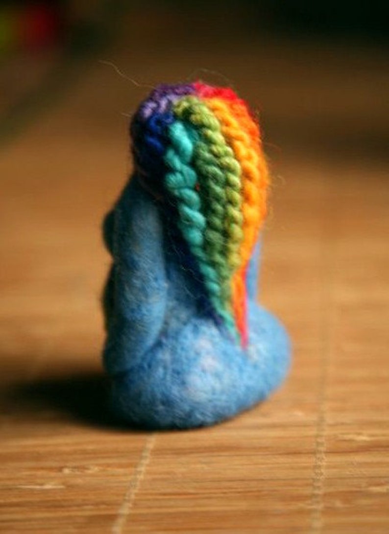 May include: A blue felt figurine with a rainbow yarn hair. The figurine is sitting on a wooden surface.