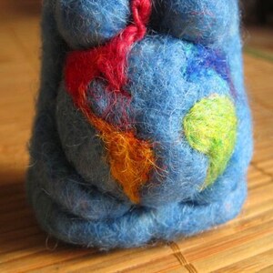 May include: A blue felt toy with colorful felt accents. The toy is shaped like a person and has a round belly with a design of colorful felt shapes.