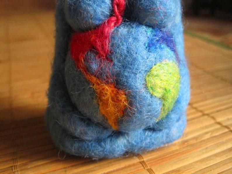 May include: A blue felt toy with colorful felt accents. The toy is shaped like a person and has a round belly with a design of colorful felt shapes.