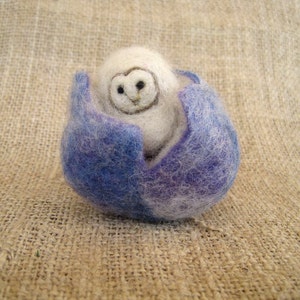 May include: A white felt owl with black eyes is nestled inside a blue felt flower.