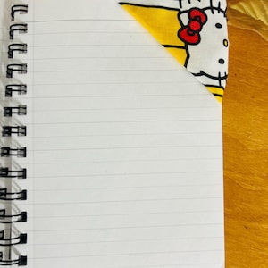 Puede incluir: Un cuaderno con espiral con papel blanco rayado. Un marcap&aacute;ginas de esquina de tela con un patr&oacute;n amarillo y blanco con un lazo rojo y detalles negros se coloca en la esquina superior derecha del cuaderno.