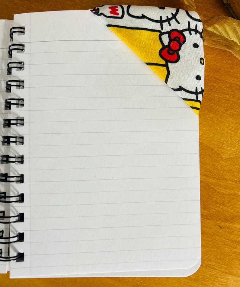 Puede incluir: Un cuaderno con espiral con papel blanco rayado. Un marcap&aacute;ginas de esquina de tela con un patr&oacute;n amarillo y blanco con un lazo rojo y detalles negros se coloca en la esquina superior derecha del cuaderno.