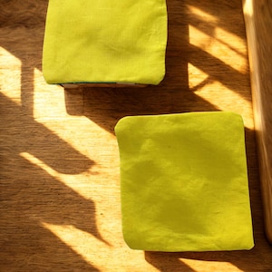Puede incluir: Dos objetos cuadrados de tela verde lima, posiblemente posavasos o peque&ntilde;os cojines, descansan sobre una superficie de madera. Los art&iacute;culos est&aacute;n hechos de una tela texturizada. La luz del sol proyecta sombras sobre la madera, creando un contraste de luz y oscuridad.