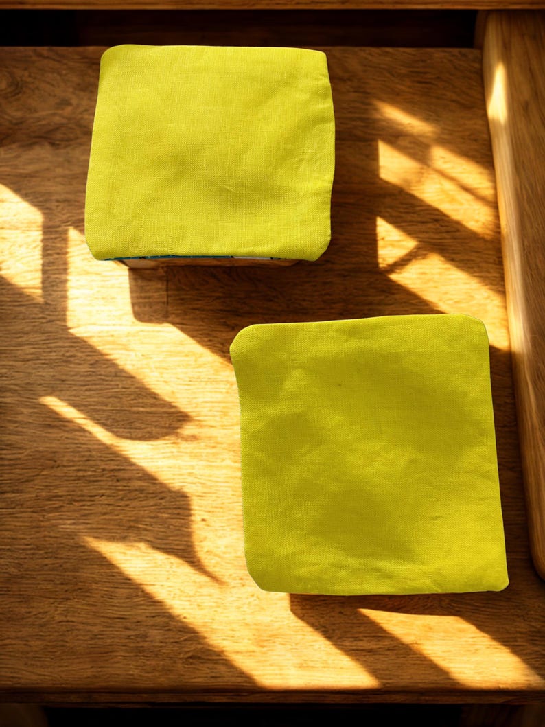 Puede incluir: Dos objetos cuadrados de tela verde lima, posiblemente posavasos o peque&ntilde;os cojines, descansan sobre una superficie de madera. Los art&iacute;culos est&aacute;n hechos de una tela texturizada. La luz del sol proyecta sombras sobre la madera, creando un contraste de luz y oscuridad.